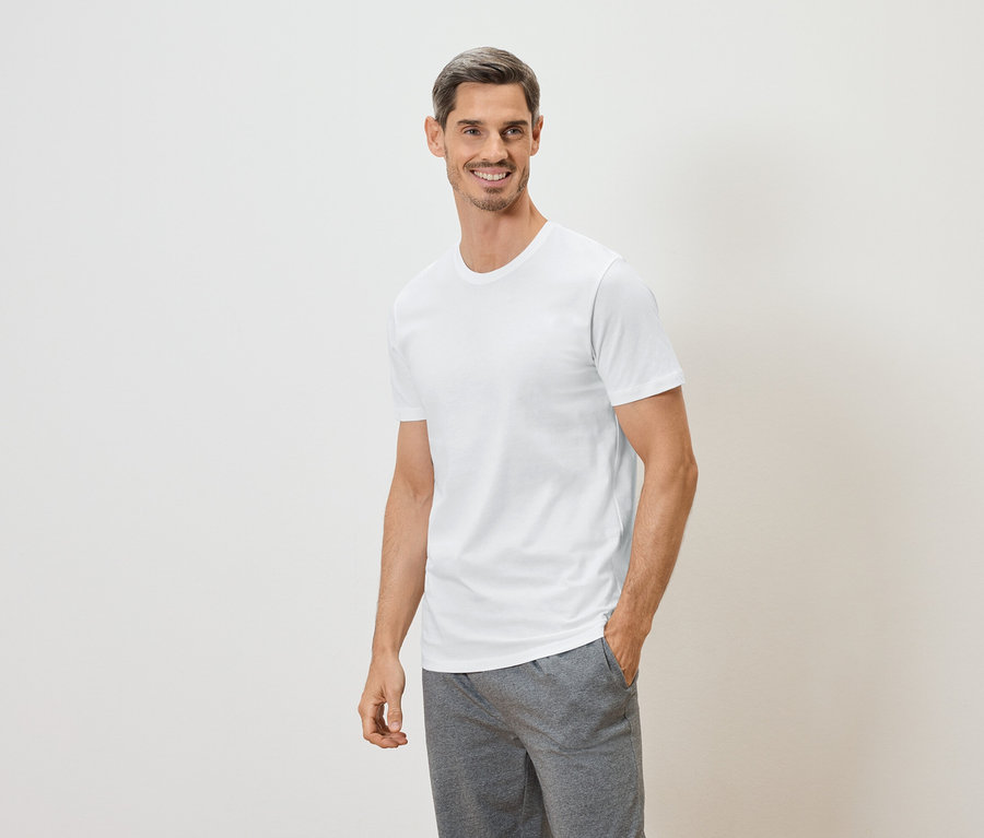 Un homme souriant, la main dans la poche, porte un t-shirt blanc et un short gris sur fond blanc.