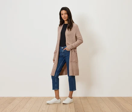 Une femme se tient devant un mur blanc, portant un long cardigan beige, un jean et des baskets blanches.