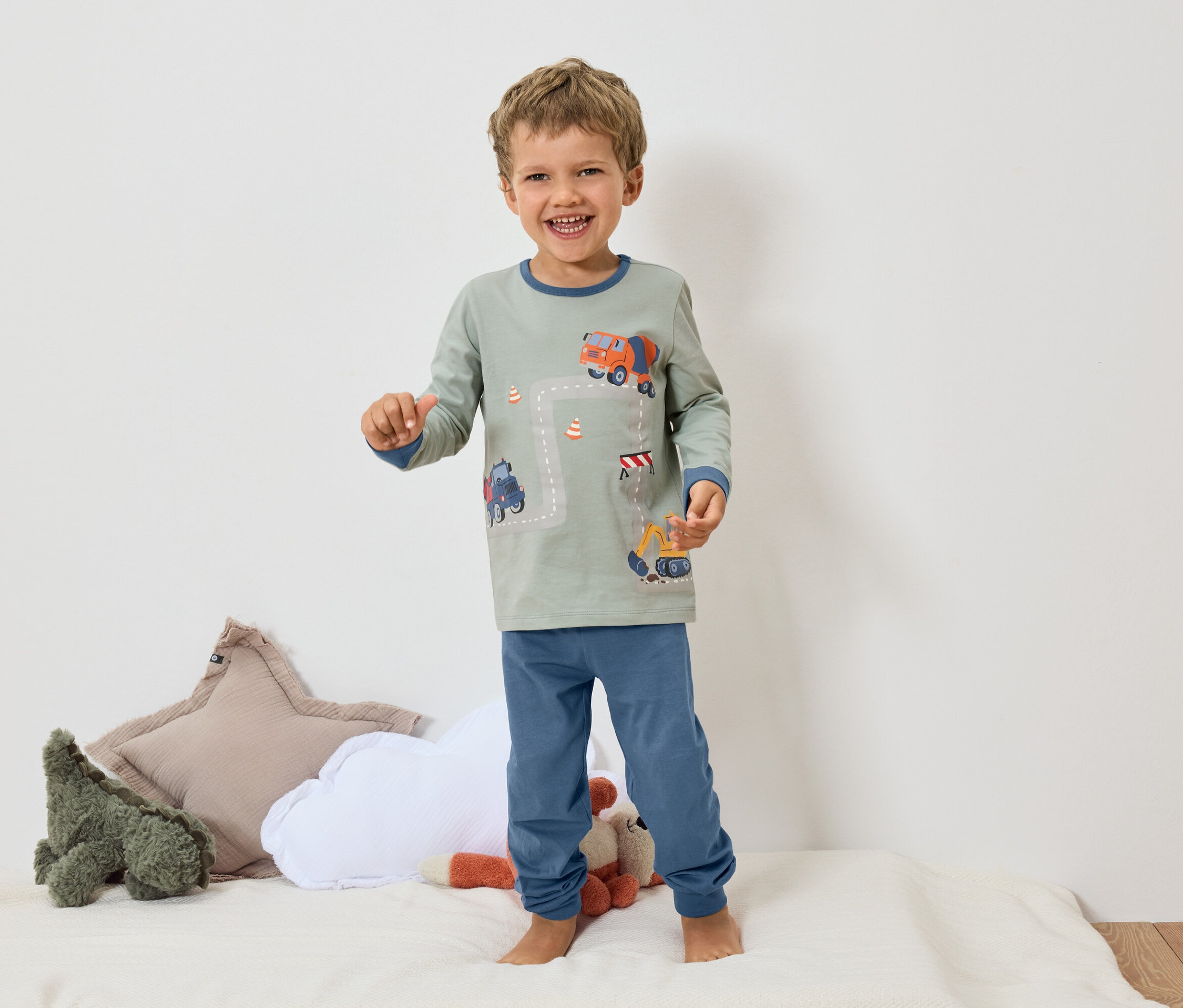 Un garçon souriant se tient pieds nus sur une surface blanche, portant un pyjama gris avec des poignets bleus et un motif d'excavatrice et de bétonnière.
