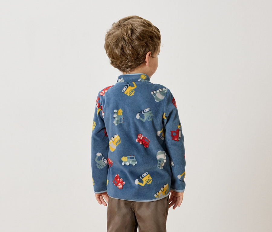 Vue arrière d'un petit garçon aux cheveux bouclés portant un pull en polaire bleu avec des motifs de véhicules colorés et un pantalon marron.