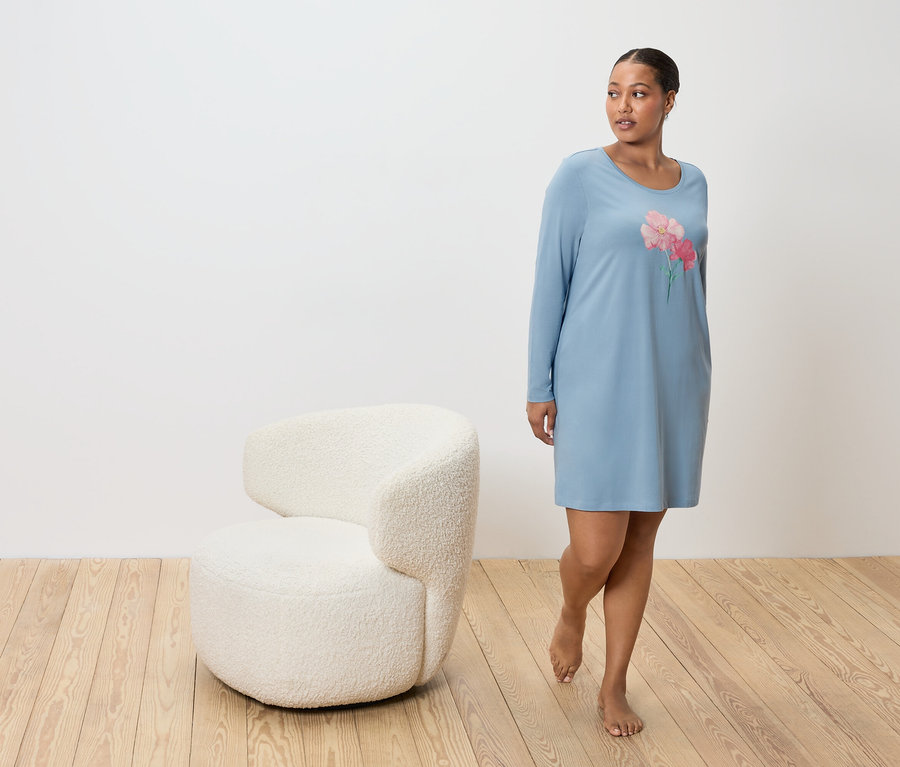 Femme en chemise de nuit bleue avec motif floral, à côté d'un fauteuil blanc sur un sol en bois.