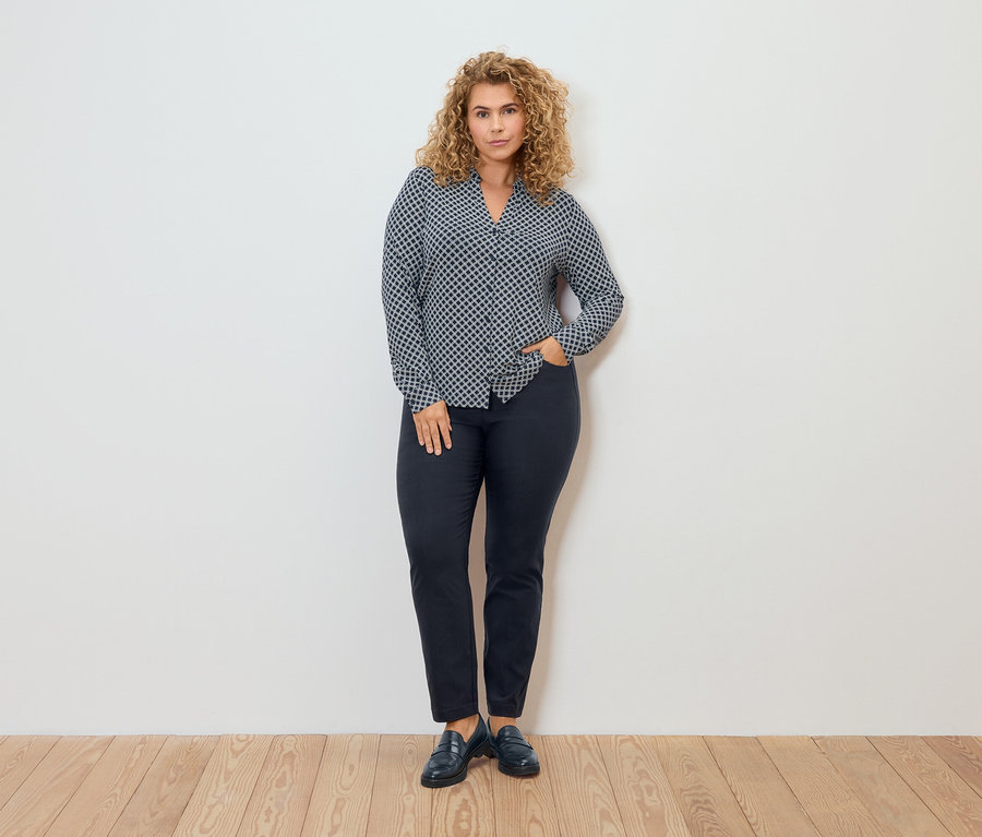 Femme en chemisier à motifs et pantalon en bengaline posant devant un mur.