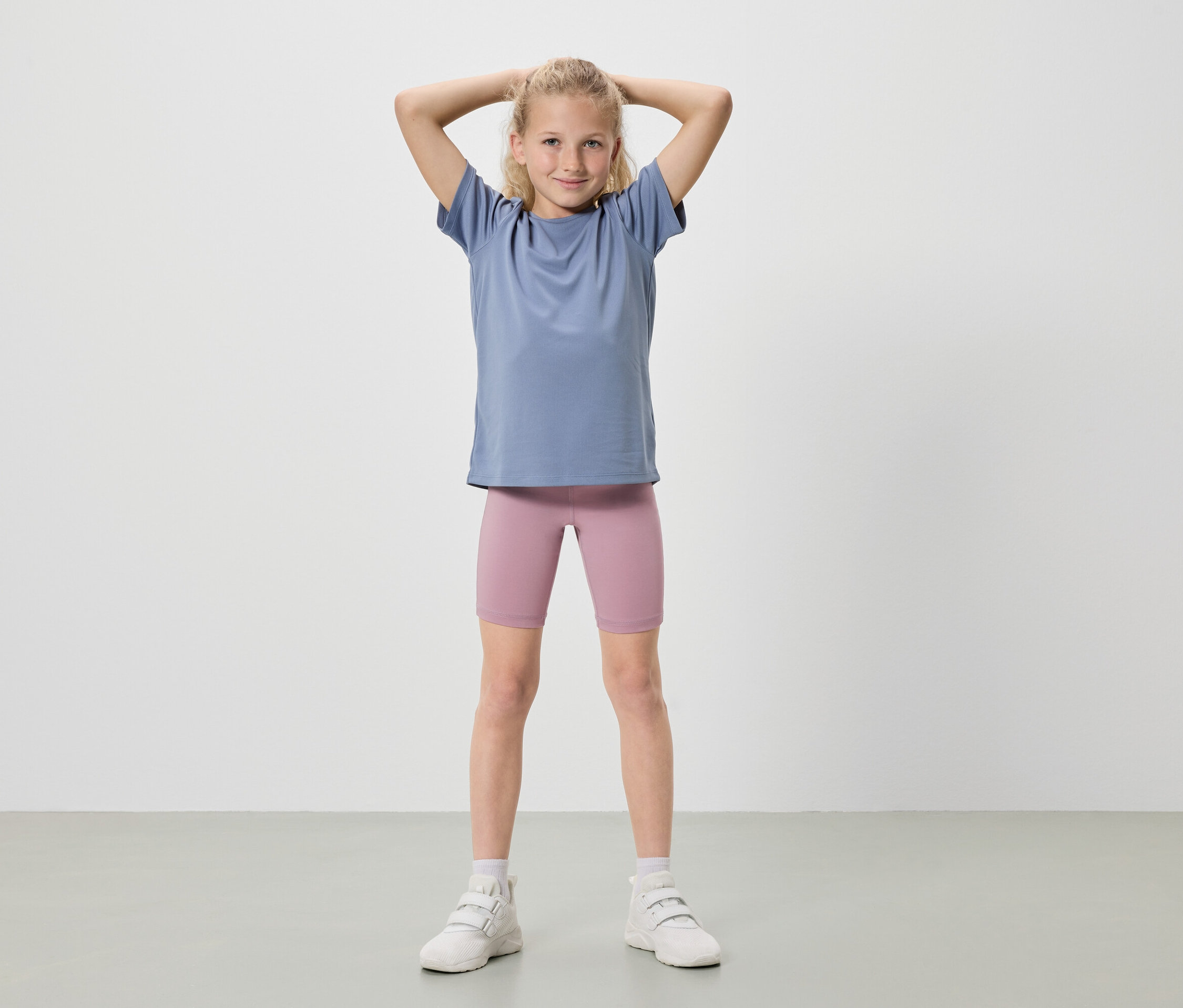 Une fille blonde se tient debout, les mains levées, portant un t-shirt bleu, un short de cycliste rose et des baskets blanches.