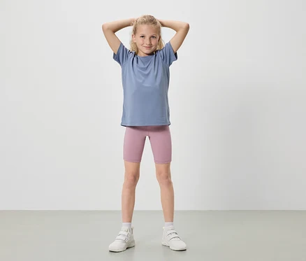 Une fille blonde se tient debout, les mains levées, portant un t-shirt bleu, un short de cycliste rose et des baskets blanches.