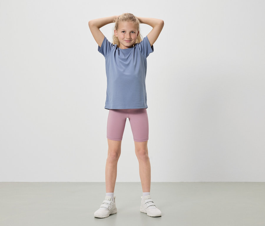 Une fille blonde se tient debout, les mains levées, portant un t-shirt bleu, un short de cycliste rose et des baskets blanches.
