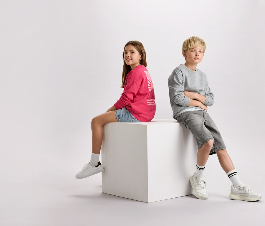 Une fille aux cheveux bruns portant un pull rose et un short en jean est assise à côté d'un garçon aux cheveux blonds portant un pull gris et un short sur un cube blanc.