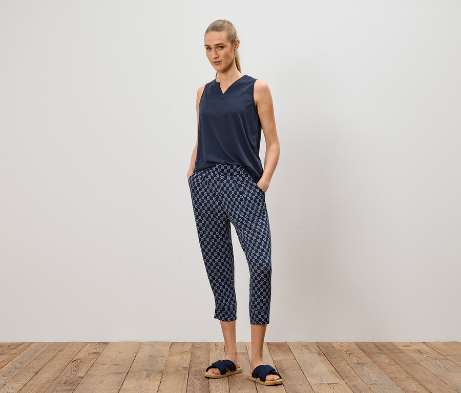 Femme debout, mains dans les poches, portant un haut bleu marine, un pantalon à motifs et des sandales bleues.