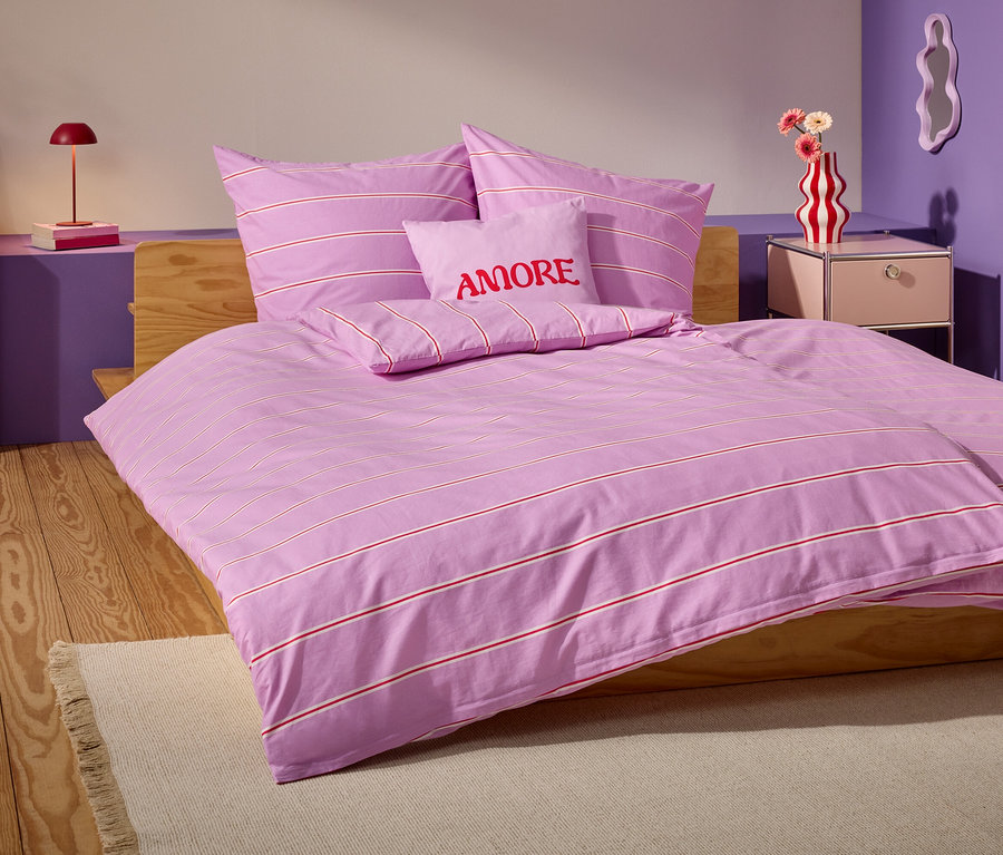 Des draps lilas rayés sont disposés sur un lit en bois avec des coussins assortis et un coussin portant l'inscription AMORE.