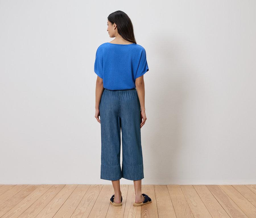 Femme vue de dos portant un chemisier bleu, une culotte en jean et des sandales devant un mur blanc.