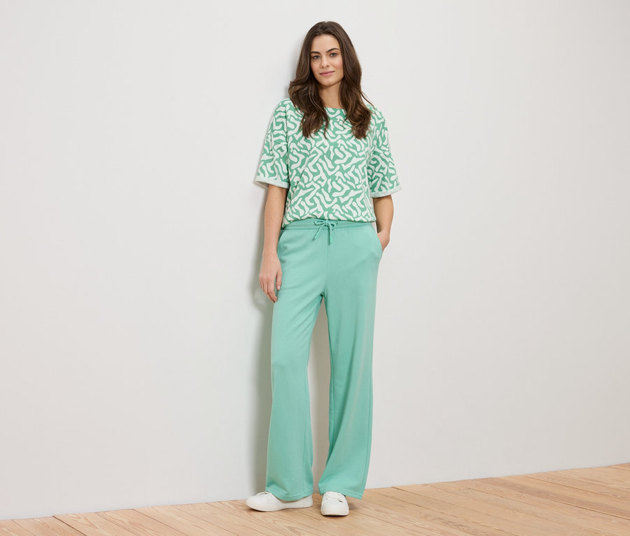 Une femme est appuyée contre un mur, portant un haut à motifs vert et un pantalon vert.