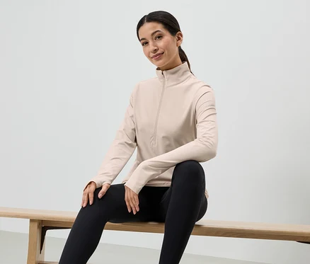 Une femme est assise sur un banc en bois, portant des leggings noirs et un haut clair à manches longues avec fermeture éclair.
