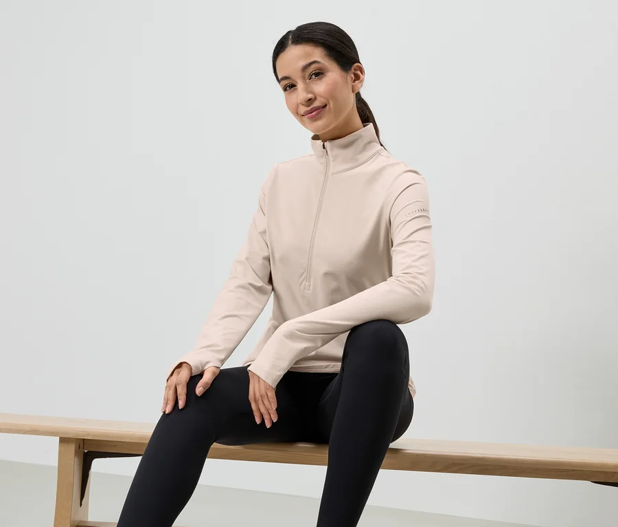 Une femme est assise sur un banc en bois, portant des leggings noirs et un haut clair à manches longues avec fermeture éclair.
