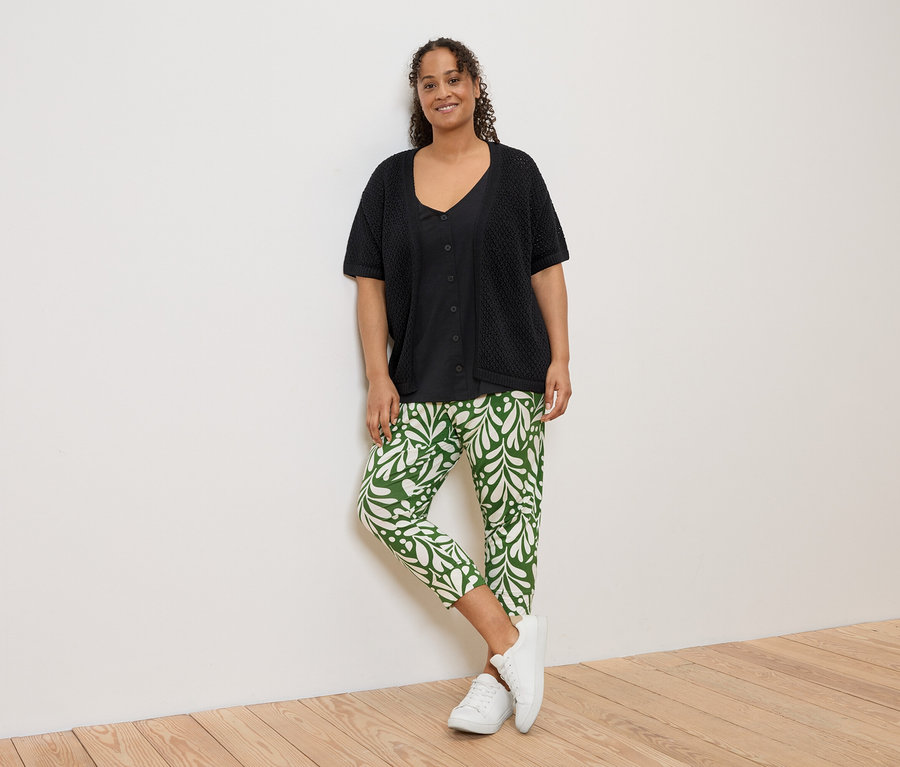 Une femme portant un cardigan noir, une chemise noire et un pantalon vert et blanc est appuyée contre un mur blanc.