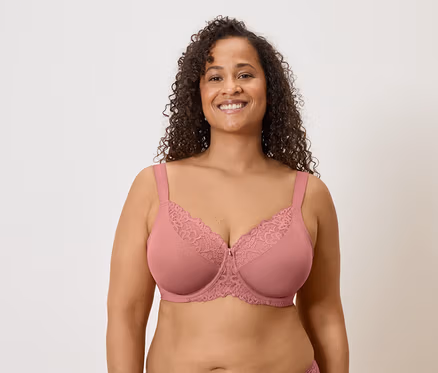 Une femme souriante aux cheveux bouclés porte un soutien-gorge rose avec de la dentelle sur un fond clair.