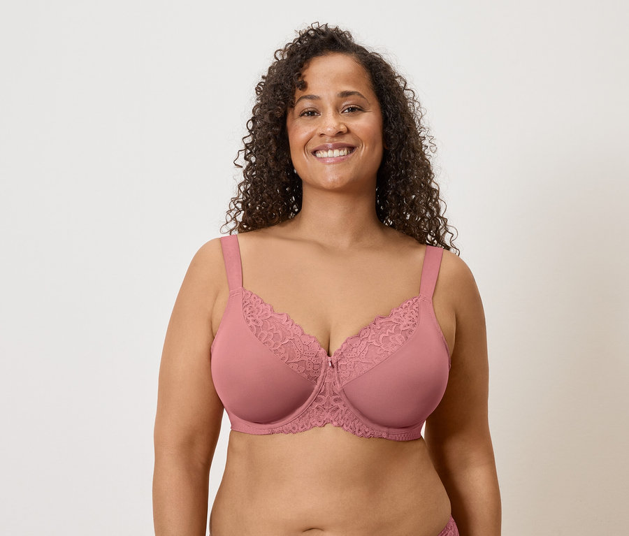 Une femme souriante aux cheveux bouclés porte un soutien-gorge rose avec de la dentelle sur un fond clair.