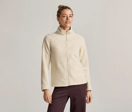 Une femme portant un chignon porte une veste en polaire crème zippée et un pantalon marron ample sur un fond clair.