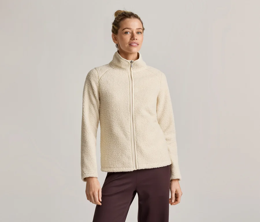 Une femme portant un chignon porte une veste en polaire crème zippée et un pantalon marron ample sur un fond clair.