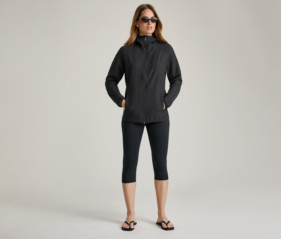 Femme portant des lunettes de soleil et une veste noire en softshell. Elle a les mains dans les poches.