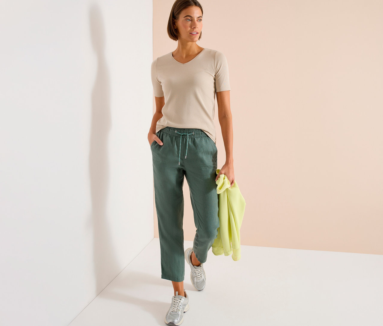 Femme debout en t-shirt beige et pantalon vert tenant un pull jaune.