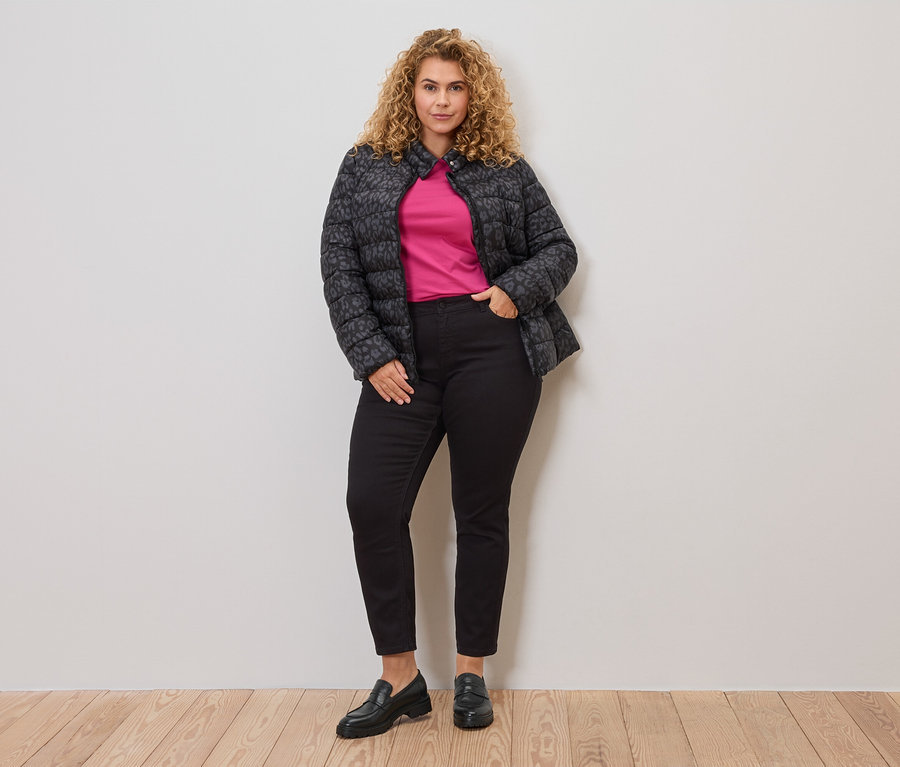 Une femme aux cheveux bouclés porte une veste matelassée gris-noir sur un haut rose, associée à un jean noir et des mocassins.