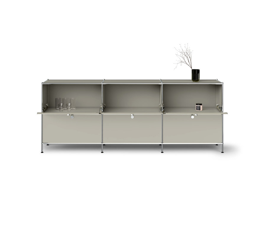 Un système d'armoires modulaires gris clair à trois sections avec des cadres chromés. Les compartiments supérieurs sont ouverts, les inférieurs ont des portes abattantes. Trois verres sont dans le compartiment gauche, une bougie rose dans le droit. Un vase noir avec une branche sèche est posé sur le dessus à droite.
