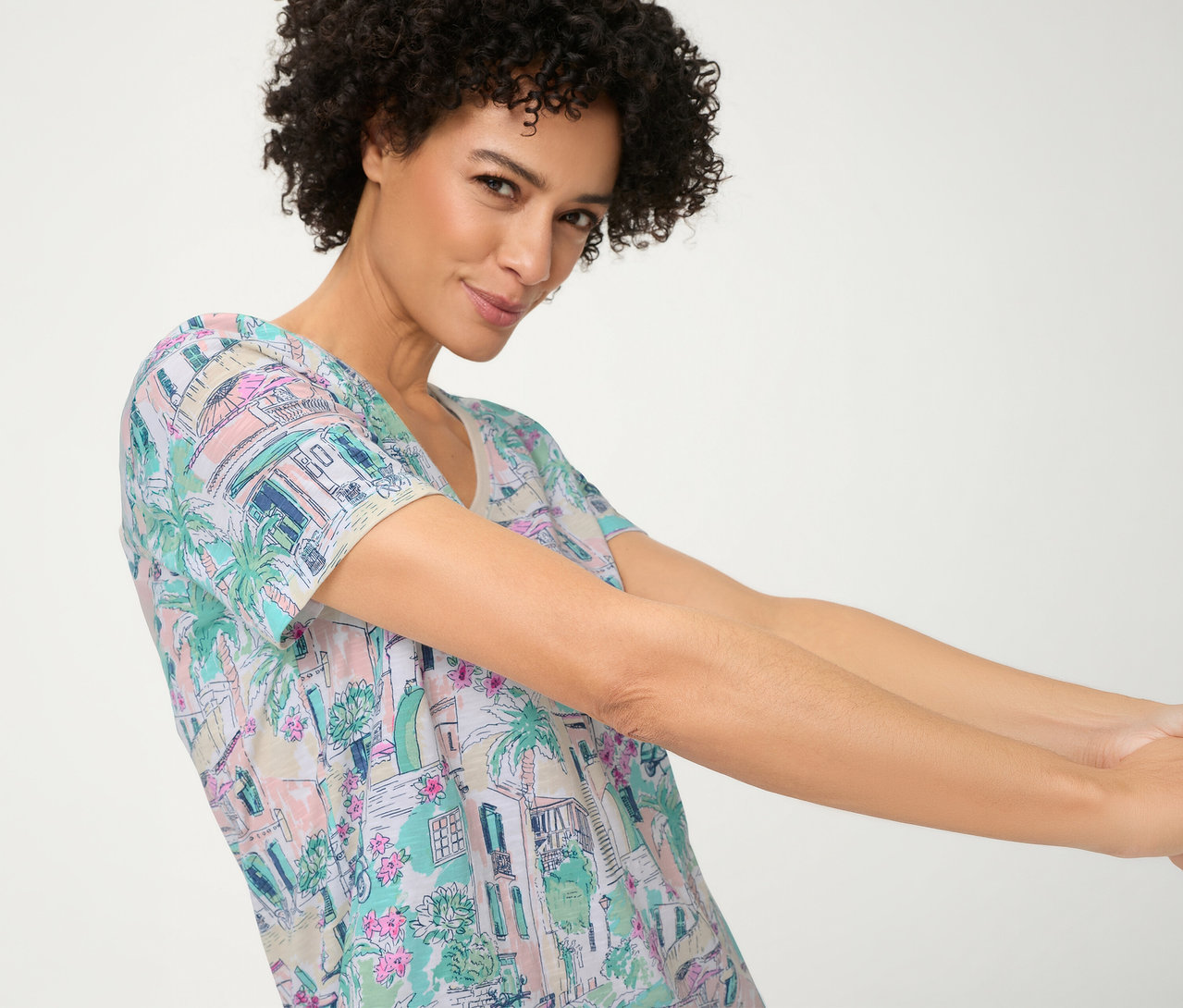 Femme aux cheveux bouclés portant un T-shirt Olsen pastel avec un motif de bâtiments, de palmiers et de fleurs, souriant et tendant les bras vers l'avant.