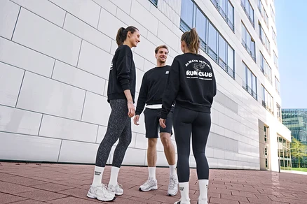 Trois personnes, deux femmes et un homme, se tiennent debout, souriantes et discutant. Tous portent des sweatshirts noirs et des chaussettes de tennis Unisex Coffee Crew blanches. L'homme porte un short de sport noir. Une femme porte des leggings de sport « ActivePower » gris-noir, l'autre des leggings de sport « ActivePower » noirs.