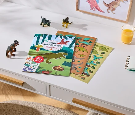 Un bureau blanc avec un livre d'autocollants de dinosaures contenant plus de 300 autocollants et deux feuilles d'autocollants. À côté se trouvent trois petites figurines de dinosaures, un verre de jus d'orange, un carnet et deux crayons de couleur. Un dessin de dinosaures encadré est accroché au mur en arrière-plan.