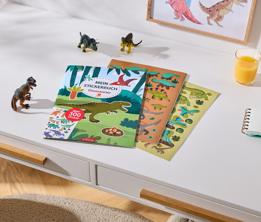 Un bureau blanc avec un livre d'autocollants de dinosaures contenant plus de 300 autocollants et deux feuilles d'autocollants. À côté se trouvent trois petites figurines de dinosaures, un verre de jus d'orange, un carnet et deux crayons de couleur. Un dessin de dinosaures encadré est accroché au mur en arrière-plan.