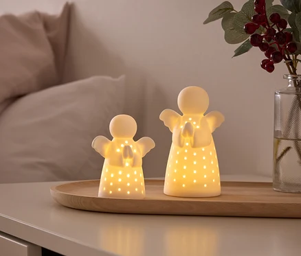 Deux figurines d'anges blanches lumineuses avec des trous découpés reposent sur un plateau en bois sur une table de chevet. À l'arrière-plan, on voit un lit et un vase avec des baies rouges.