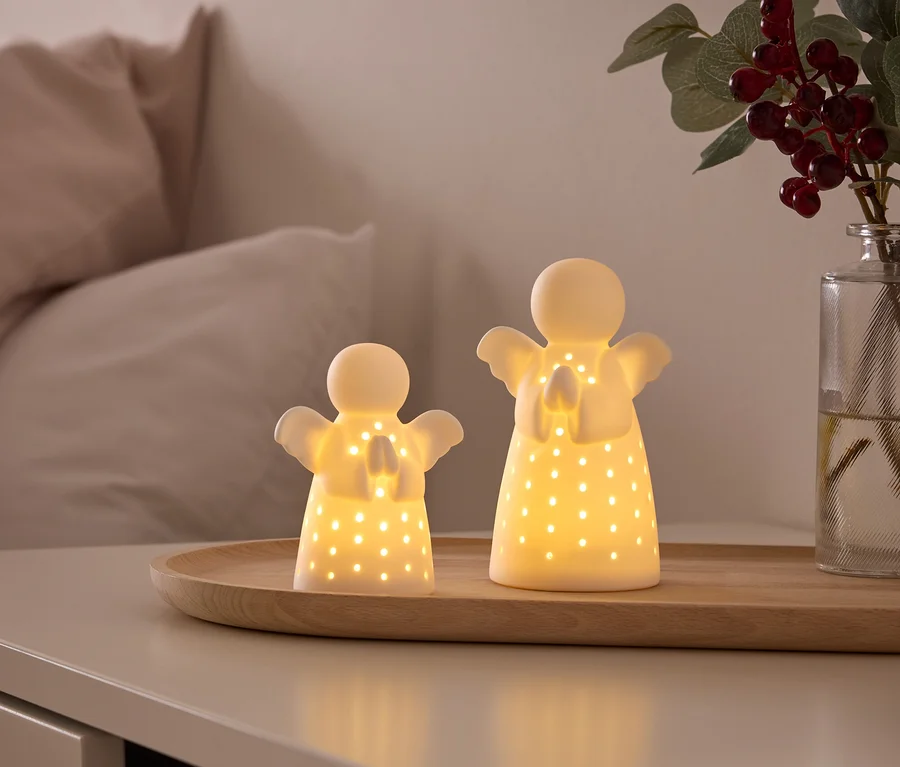 Deux figurines d'anges blanches lumineuses avec des trous découpés reposent sur un plateau en bois sur une table de chevet. À l'arrière-plan, on voit un lit et un vase avec des baies rouges.