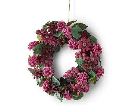 Couronne florale composée de fleurs violettes et bordeaux et de feuilles vertes, suspendue par une ficelle sur fond blanc.