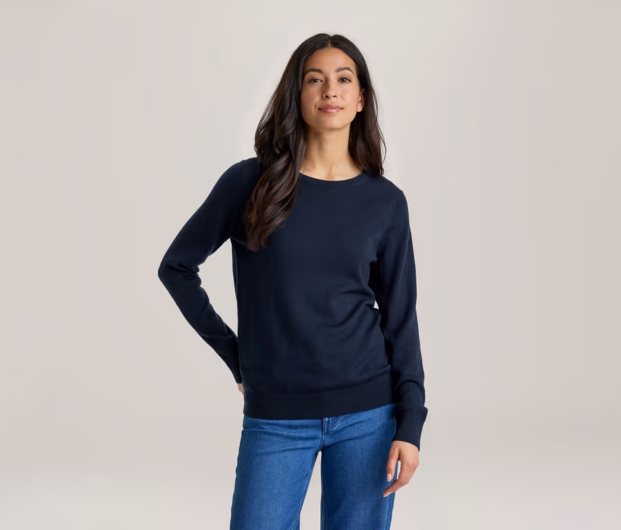 Une femme aux longs cheveux foncés, vêtue d'un haut bleu foncé à manches longues et d'un jean bleu, se tient la main sur la hanche, regardant devant elle.