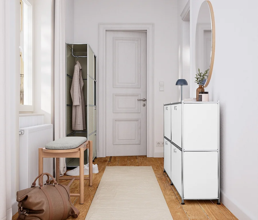 Couloir lumineux avec parquet et tapis beige. À gauche, un banc en bois massif avec rembourrage et espace de rangement est placé sous une fenêtre, à côté d'un sac de voyage marron et d'une armoire ouverte. À droite, un buffet blanc surmonté d'un miroir rond.
