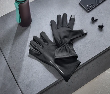 Une paire de gants noirs avec des points réfléchissants et des bouts de doigts compatibles avec les écrans tactiles, une gourde foncée avec une sangle turquoise, un smartphone noir et des écouteurs sans fil noirs sont disposés sur une surface grise.