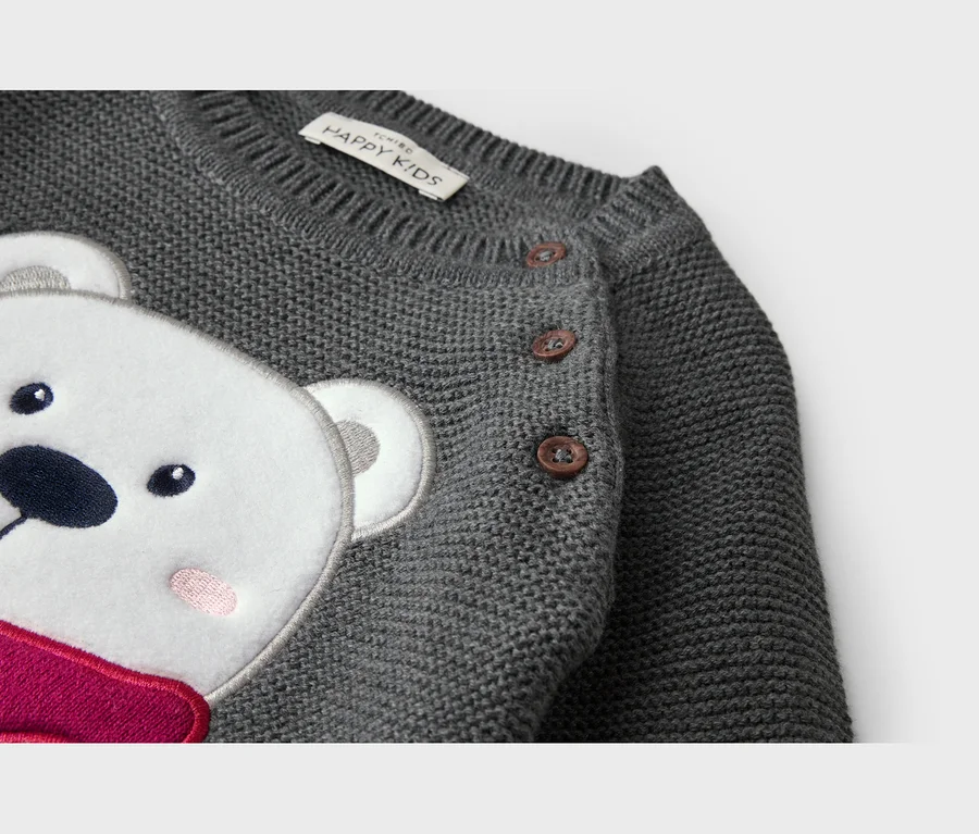 Gros plan d'un pull enfant en tricot gris avec une application d'ours polaire blanc aux yeux noirs, nez, joues roses et écharpe rose. Trois boutons marron sont visibles sur l'épaule et une étiquette "HAPPY KIDS" près de l'encolure.