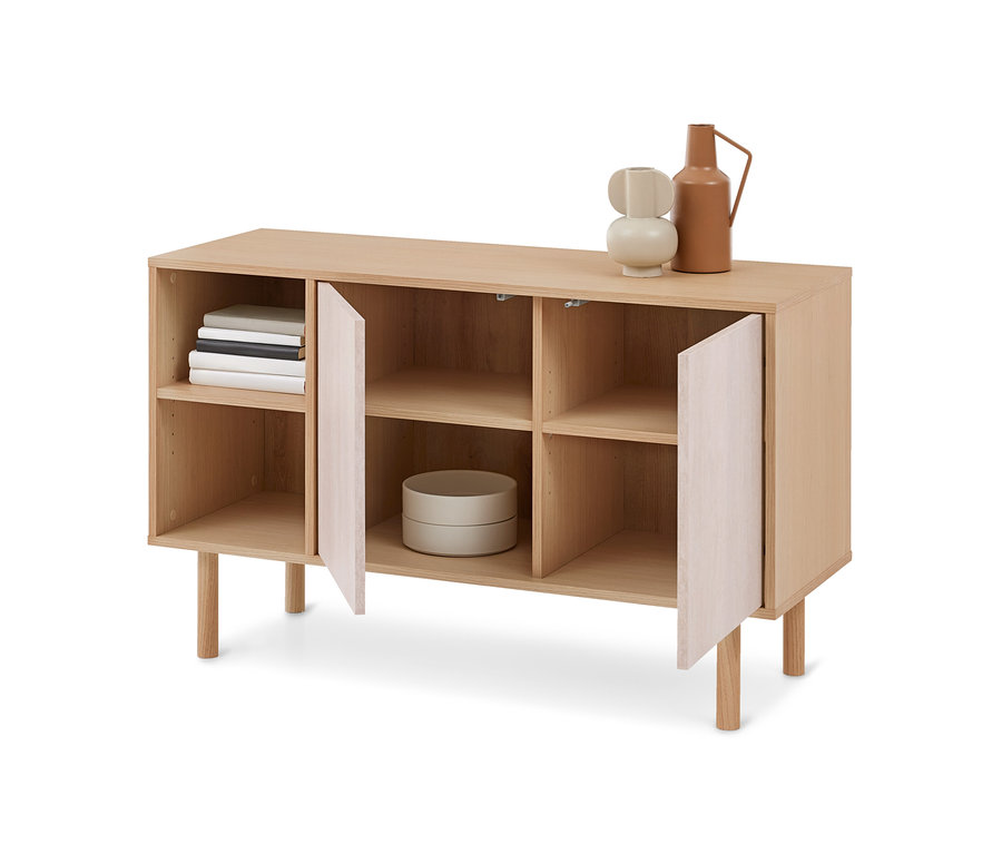 Un buffet en bois clair avec des étagères ouvertes et deux portes légèrement entrouvertes. Des livres sont posés sur une étagère supérieure gauche, et un récipient gris clair sur une étagère inférieure centrale. Un vase abstrait beige et une cruche en terre cuite sont posés sur le dessus.