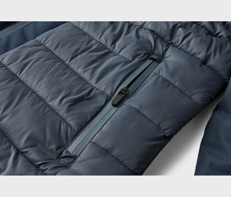 Gros plan d'une veste matelassée bleu foncé avec des coutures horizontales et une poche zippée diagonale. La fermeture éclair a une tirette noire et une bande plus claire.