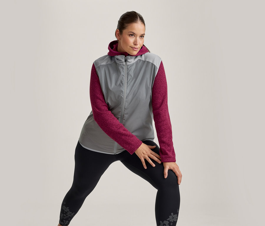 Femme en veste à capuche grise et magenta et leggings noirs à motifs s'étirant, un genou plié vers l'avant.