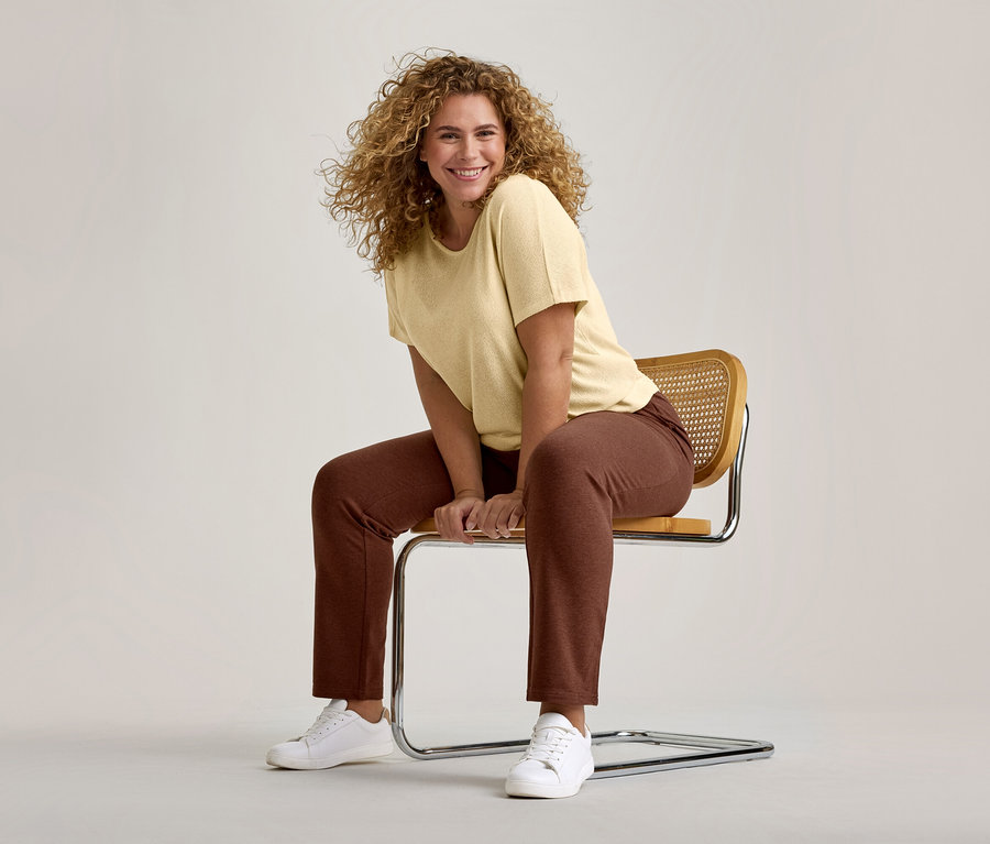 Femme souriante aux cheveux bouclés, vêtue d'un t-shirt jaune clair, d'un pantalon marron et de baskets blanches, assise sur une chaise moderne.