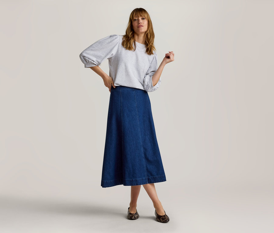 Femme debout, portant un sweat-shirt gris clair à manches bouffantes, une jupe midi en jean bleu foncé et des chaussures plates foncées.