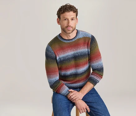 Homme assis sur un tabouret, portant un pull à col rond rayé coloré dans des tons de rouge, bleu, vert et marron, et un jean bleu.