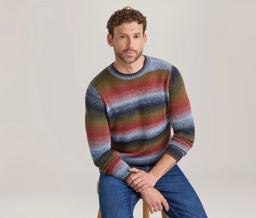 Homme assis sur un tabouret, portant un pull à col rond rayé coloré dans des tons de rouge, bleu, vert et marron, et un jean bleu.