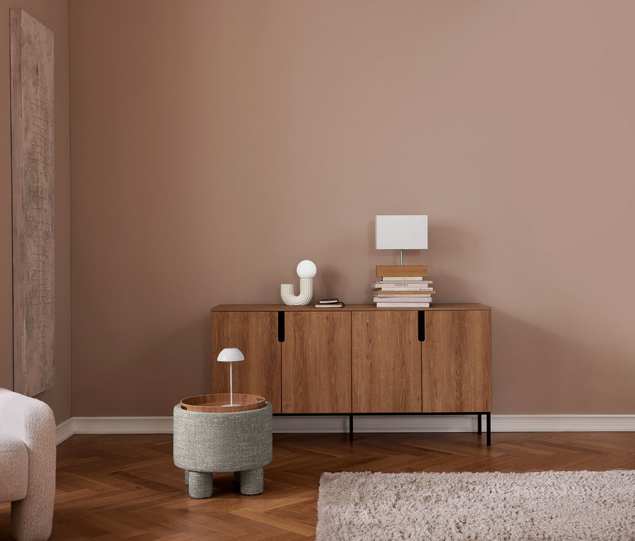 Une pièce moderne avec des murs marron clair et un parquet en chevrons. Un fauteuil de couleur claire est partiellement visible sur la gauche. Au centre, un pouf rembourré gris avec un plateau en bois accueille une petite lampe de table blanche. Derrière, un buffet en bois à quatre portes expose un objet décoratif blanc, une petite pile de livres et une lampe de table blanche rectangulaire sur une autre pile de livres. Un tapis shaggy gris clair est posé sur le sol.