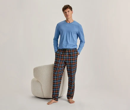 Un homme debout, portant un t-shirt bleu à manches longues et un pantalon de pyjama à carreaux rouges, verts et bleus. Les mains dans les poches, pieds nus. Un fauteuil clair et texturé est derrière lui.
