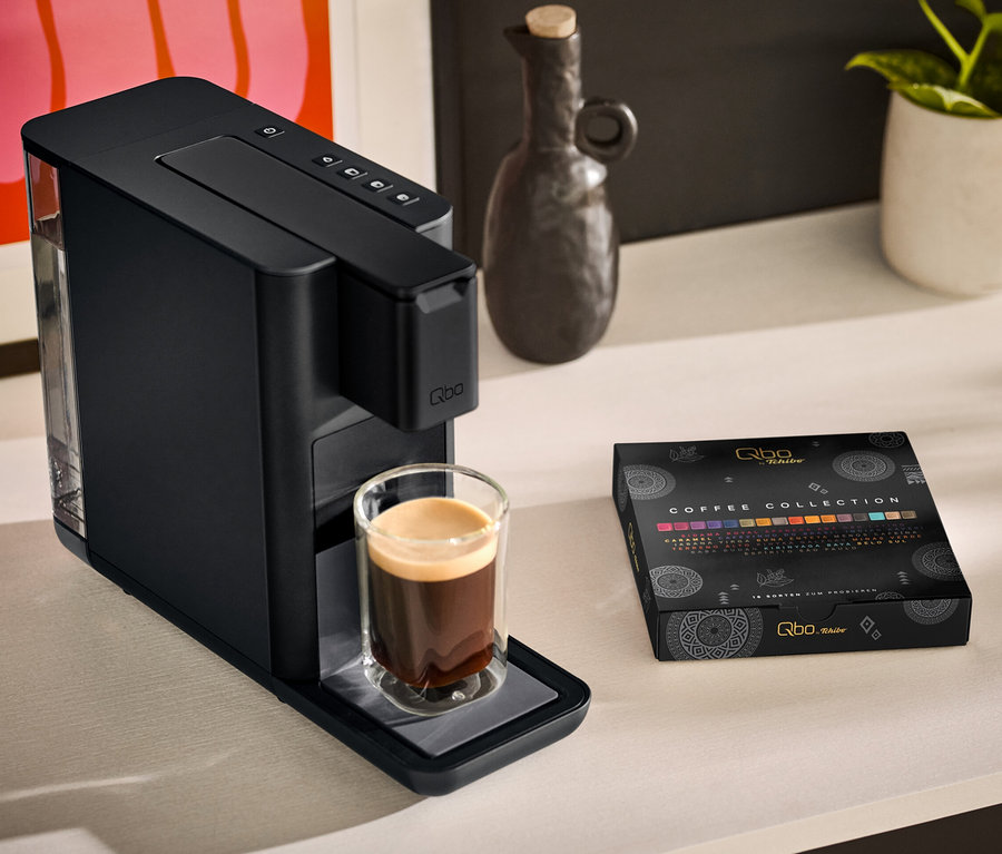 Une machine à café Qbo ICONIC Dark Chrome avec une tasse de café à côté et un paquet de Qbo Collection - 16 capsules sur une table.