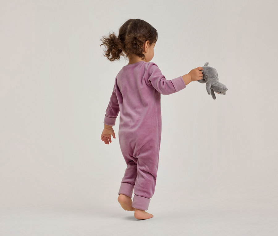 Un jeune enfant, vu de dos, marche pieds nus dans une combinaison en velours violette et tient un animal en peluche gris dans sa main droite.