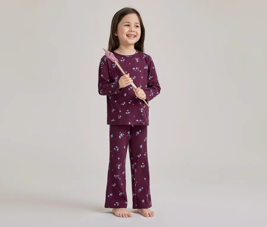 Une jeune fille souriante est debout pieds nus, vêtue d'un pyjama côtelé bordeaux à motif floral clair, tenant une baguette magique jouet rose et marron.