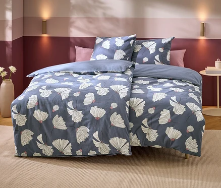 Un lit avec une parure de lit bleue à motifs floraux blancs et accents roses. Deux oreillers roses sont derrière. Le mur est bicolore violet, avec deux appliques murales dorées. À gauche, un vase avec des fleurs blanches, à droite, une table d'appoint avec des livres.