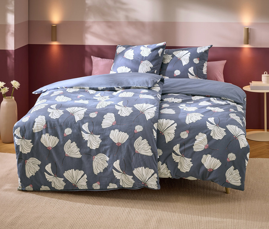 Un lit avec une parure de lit bleue à motifs floraux blancs et accents roses. Deux oreillers roses sont derrière. Le mur est bicolore violet, avec deux appliques murales dorées. À gauche, un vase avec des fleurs blanches, à droite, une table d'appoint avec des livres.